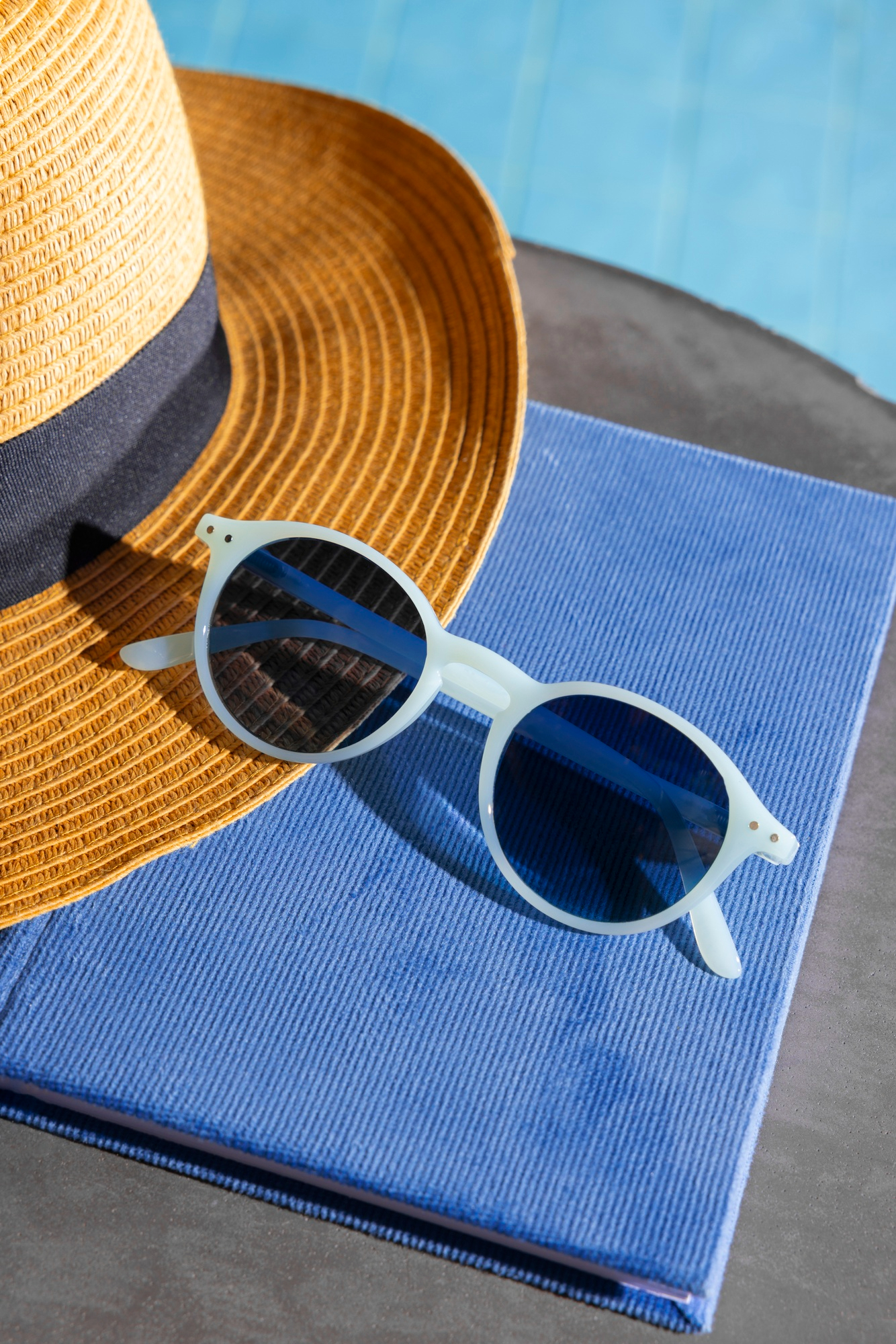 Round white-framed sunglasses resting on a blue cloth next to a straw hat with a black band near a pool.