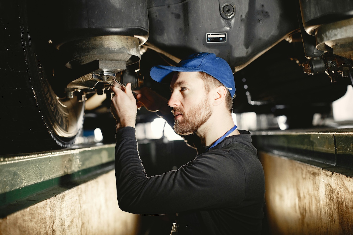Man lying on ground working under a car elevated on a metal stand, with a can of WD-40 and tools nearby.