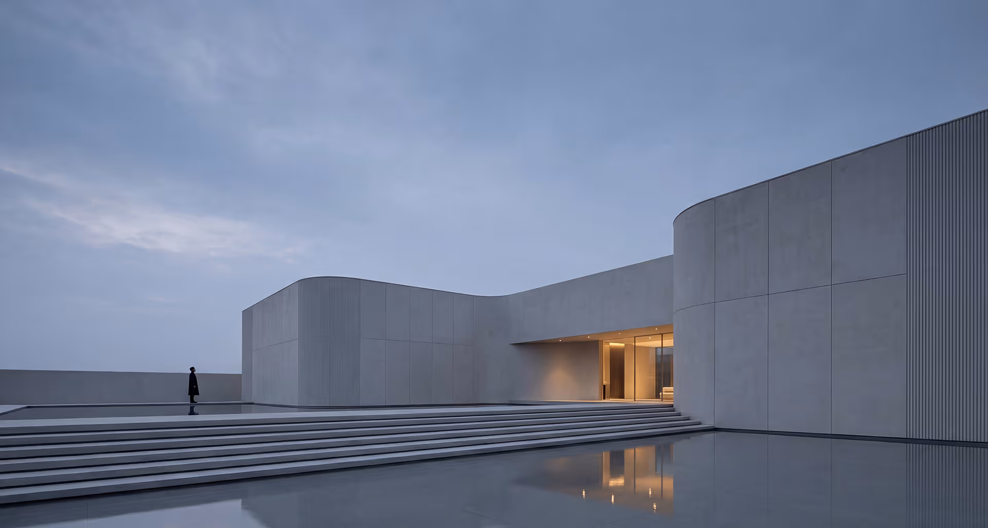 Modern minimalist building with curved concrete walls, broad steps, and shallow reflective pool at dusk with a person standing near the pool edge.