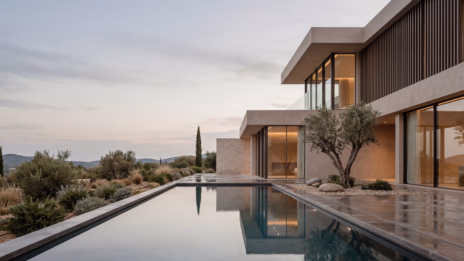 Modern house with large glass windows and a sleek rectangular pool reflecting the building and surrounding landscape at dusk.