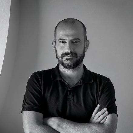 Portrait of a bald man with a beard wearing a black polo shirt, standing with arms crossed against a plain wall.