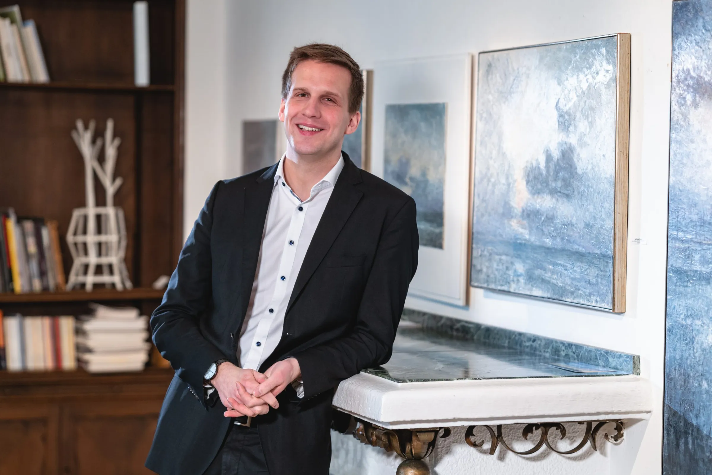 Ein lächelnder Mann im schwarzen Anzug lehnt an einem Kamin mit blau-grauen Gemälden im Hintergrund.