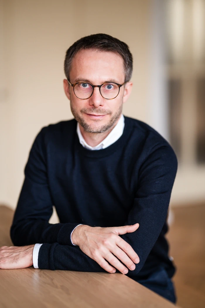 Mann mit dunklem Pullover und Brille sitzt mit verschränkten Armen an einem Holztisch vor uns.