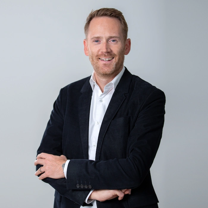 Ein lächelnder Mann mit kurzen blonden Haaren trägt ein weißes Hemd und eine schwarze Jacke und steht vor einem neutralen grauen Hintergrund.