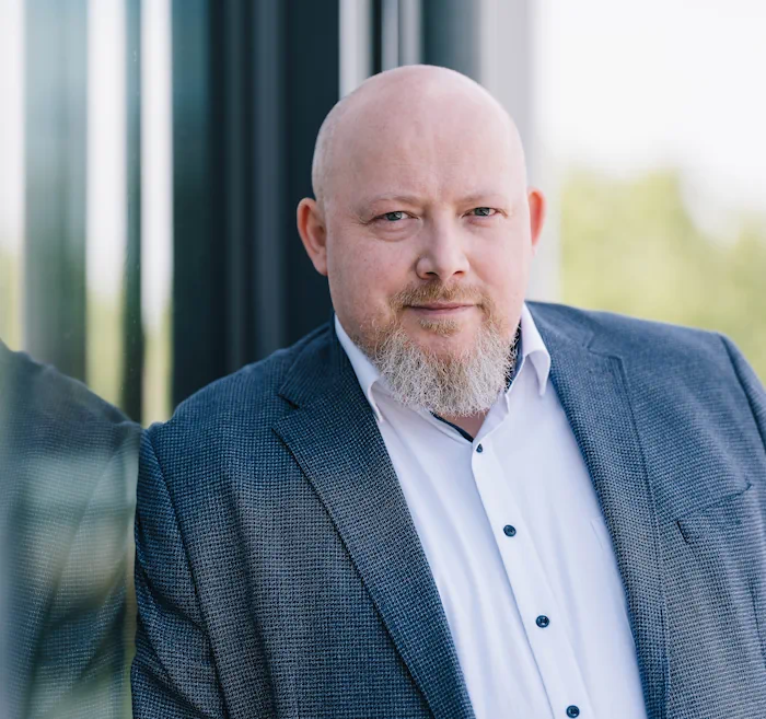 Porträt eines lächelnden glatzköpfigen Mannes mit Bart in einem grauen Sakko und weißem Hemd vor einem Gebäude mit Glasfenstern.