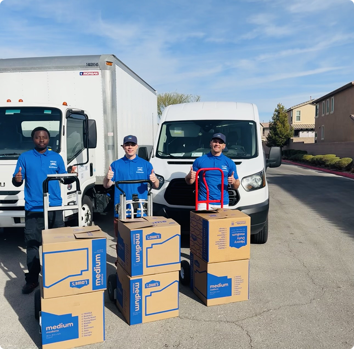 Three movers in blue uniforms giving thumbs up, standing behind dollies loaded with stacked medium moving boxes, in front of two white moving trucks on a sunny residential street.
