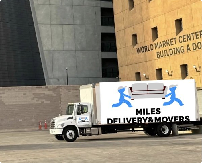 A white delivery truck with a sign showing two blue figures carrying a sofa and text reading 'MILES DELIVERY & MOVERS' parked outside a building labeled 'WORLD MARKET CENTER BUILDING A'.