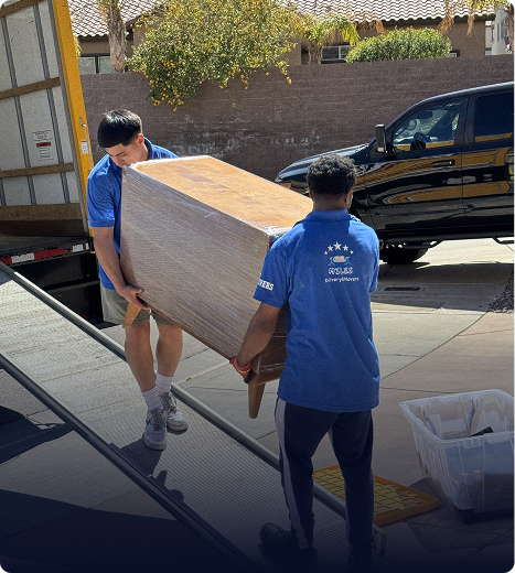 Two movers in blue uniforms carrying a large wrapped piece of furniture up a ramp to a moving truck.