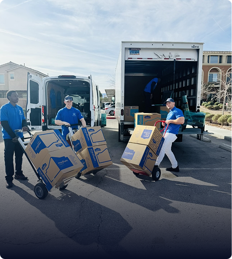 Three men in blue shirts unloading cardboard boxes from two trucks using hand trucks in a residential area.