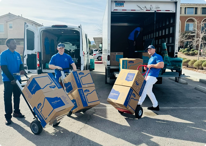 Three movers in blue shirts using dollies to transport medium-sized moving boxes in front of a truck and van in a suburban neighborhood.