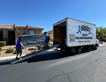 Two movers carrying a large wrapped piece of furniture up a ramp into a delivery truck on a sunny residential street.