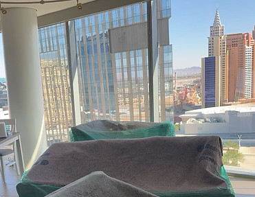 Indoor view of furniture covered in protective blankets near large windows overlooking city buildings and a clear sky.