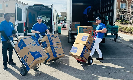 Three men in blue shirts loading large cardboard boxes onto trucks using hand trucks in a parking lot.