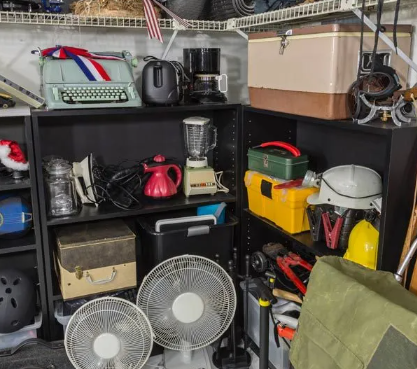Black shelving unit filled with various household items including a green typewriter, coffee makers, blender, fans, and a cooler in a cluttered storage space.