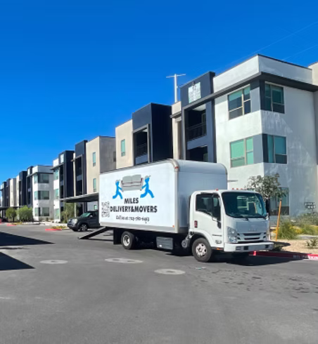 A white delivery truck labeled 'Miles Delivery & Movers' parked on a residential street with modern apartment buildings and a clear blue sky in the background.