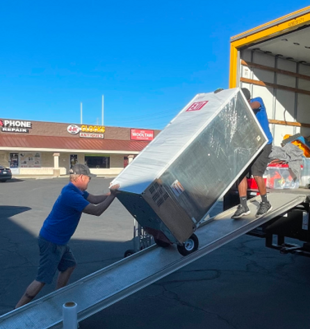 Two men loading a large boxed item on a dolly up a metal ramp into a yellow moving truck in a parking lot.
