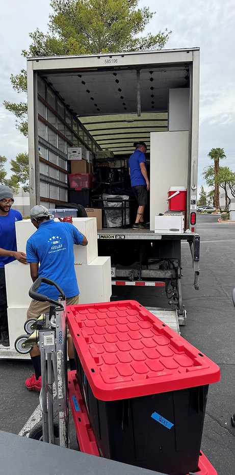 Two men in blue shirts unloading white boxes from the back of a truck parked outdoors with a red and black plastic container in the foreground.