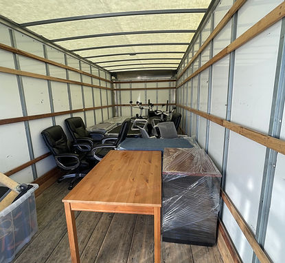 Interior of a moving truck loaded with office furniture including chairs, desks, and filing cabinets, some wrapped in plastic.