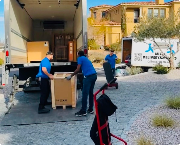 Two movers loading a large wooden crate into a truck outside a house, with another mover behind and a hand truck in the foreground.