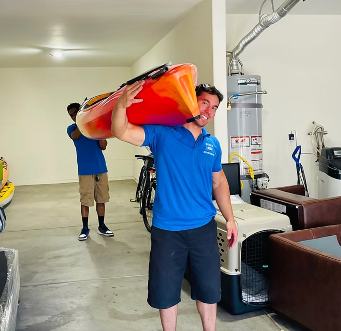 Two men in blue shirts carrying a red and orange kayak indoors near bikes and household items.
