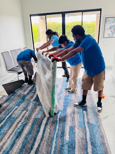 Four people in blue shirts moving a large wrapped mattress or piece of furniture on a striped blue and gray rug in a room with a glass sliding door.