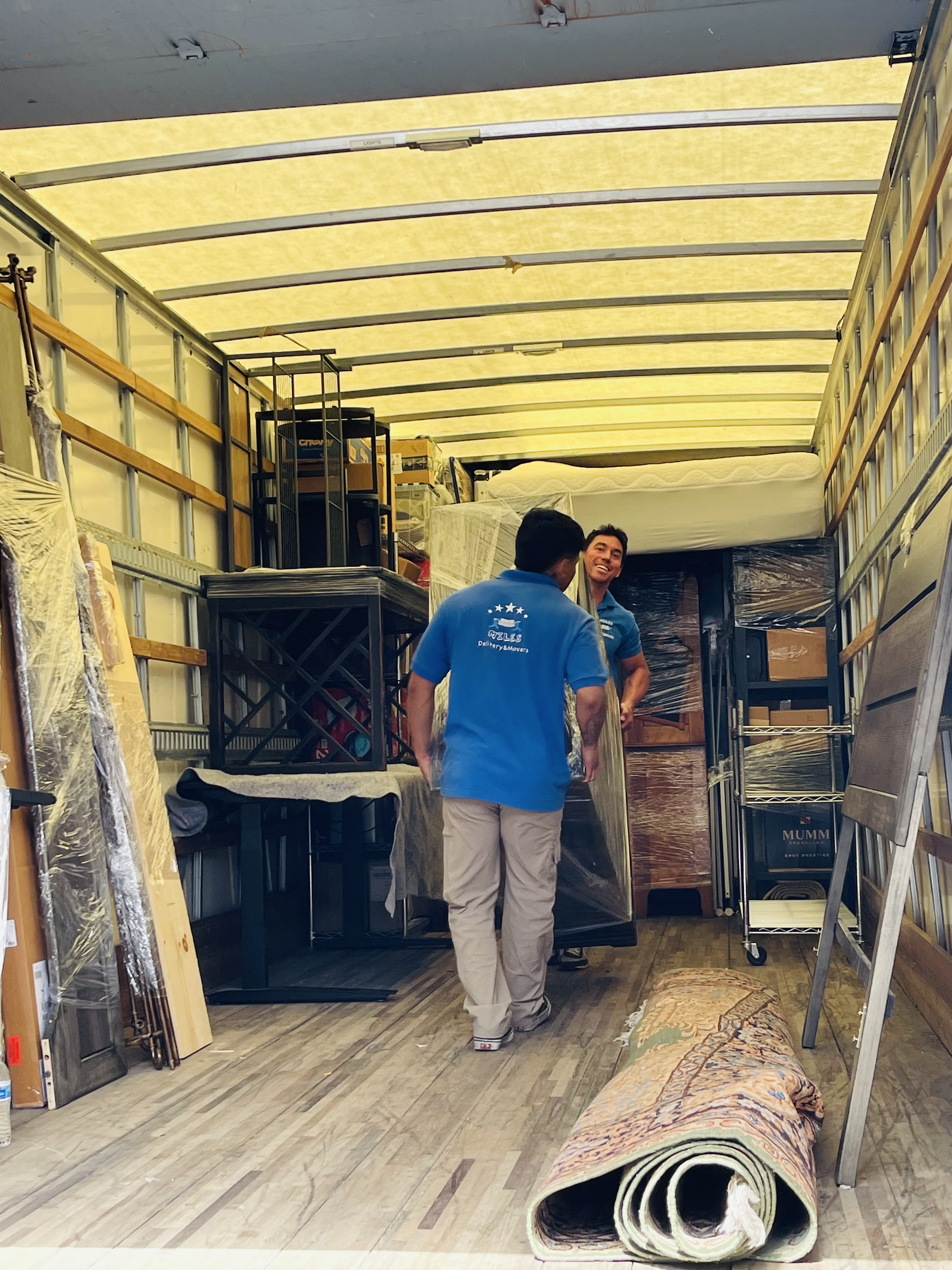 Three workers in blue shirts using hand trucks to move stacked boxes from a white van and truck in a residential area.
