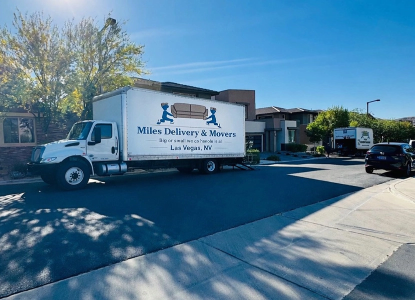 A white delivery truck labeled 'Miles Delivery & Movers' parked on a residential street with modern apartment buildings and a clear blue sky in the background.