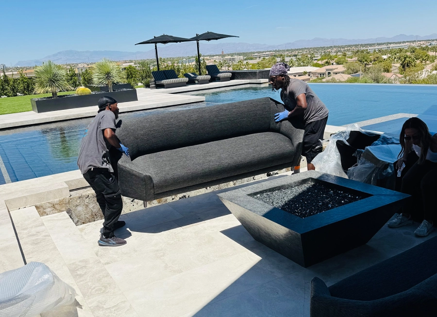 Three movers wearing blue shirts unloading a large black container from a truck in a suburban neighborhood under a clear blue sky.