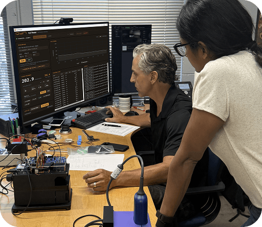 Two engineers work at a computer terminal, examining data and a hardware setup, showcasing their collaborative technical efforts.
