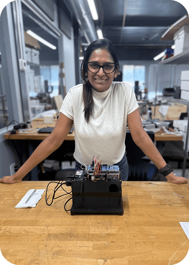 A female engineer leaning on a wooden workbench, smiling alongside a prototype device with exposed wiring in a workshop.