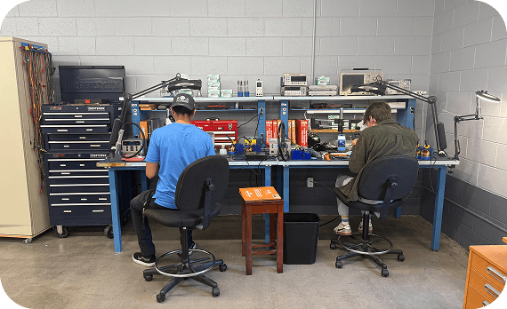 Two technicians sitting at workstations in an electronics workshop, working with equipment and tools.
