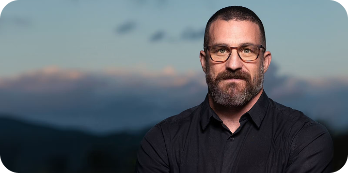A headshot of Andrew Huberman wearing glasses and a black button-up shirt, looking directly at the camera outdoors against a soft-focus dusk sky.