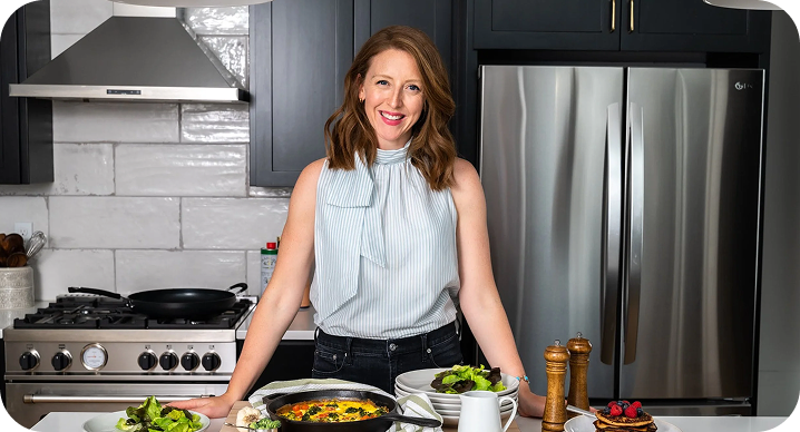 A smiling woman with shoulder-length reddish-brown hair standing in a modern kitchen with stainless steel appliances, wearing a sleeveless blue-and-white striped high-neck top, leaning over a counter with freshly prepared food, including a frittata, salad, and pancakes.