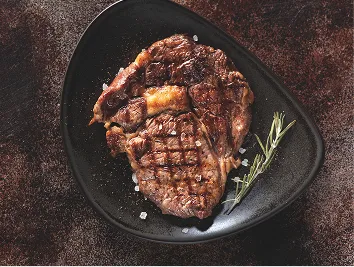 Gegrilltes Ribeye-Steak mit Salz und Rosmarin auf einem schwarzen dreieckigen Teller.
