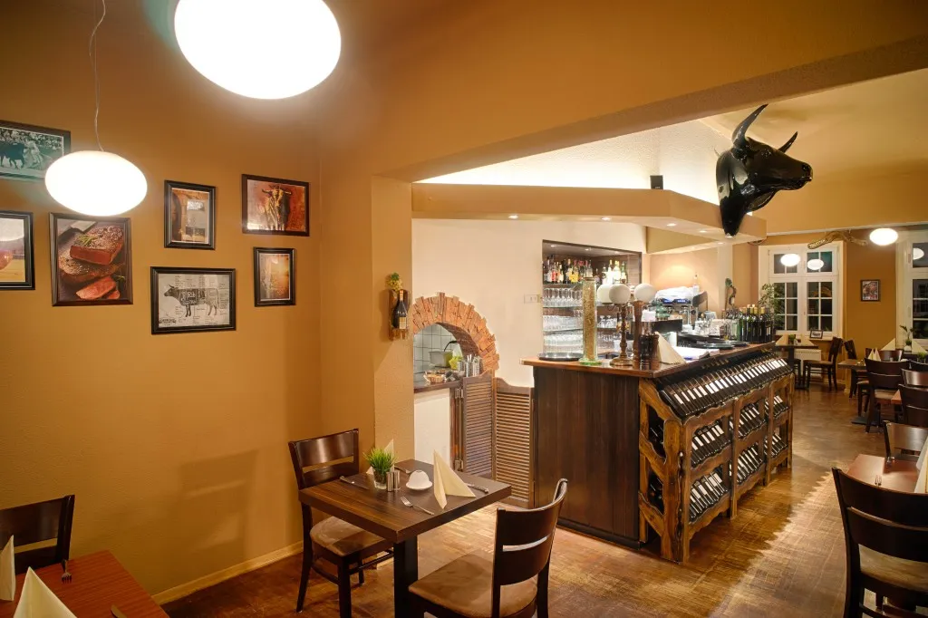 Gemütliches Restaurant-Interieur mit Holztischen und Stühlen, einer mit Weinflaschen bestückten Bar, gerahmten Bildern an einer hellbraunen Wand, zwei hängenden weißen Pendelleuchten und einem an der Decke befestigten schwarzen Stierkopf.