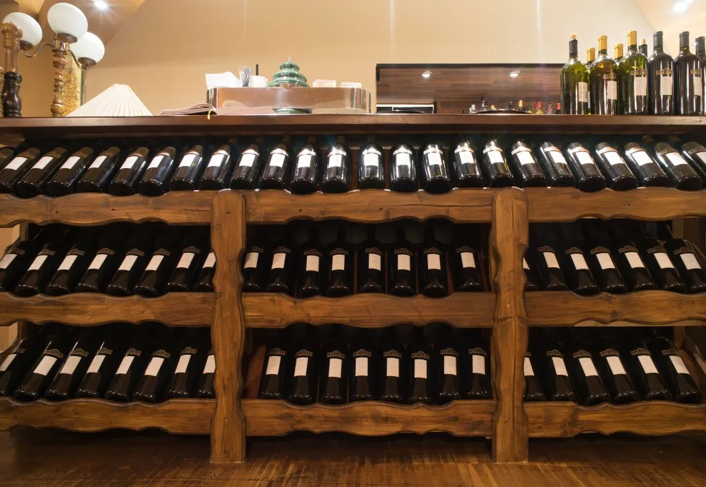 Ein hölzernes Weinregal, gefüllt mit mehreren Reihen dunkler Weinflaschen mit weißen Etiketten in einem warm beleuchteten Raum.