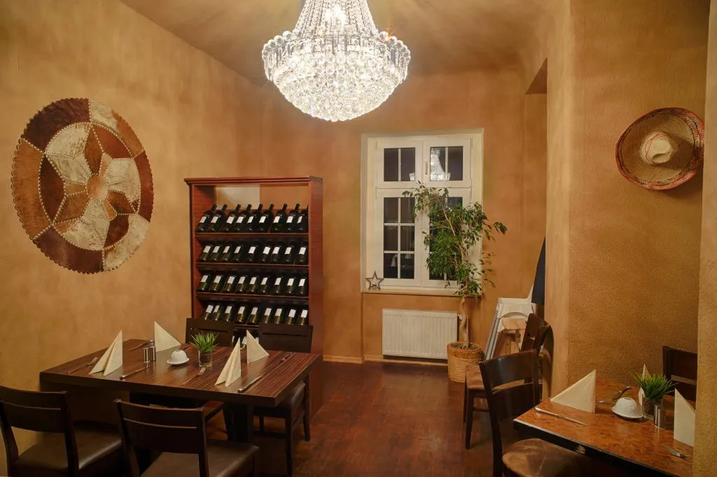 Gemütliches Restaurant-Interieur mit Holztischen, die mit gefalteten Servietten eingedeckt sind, Weinregal, Topfpflanze und Kristallleuchter.