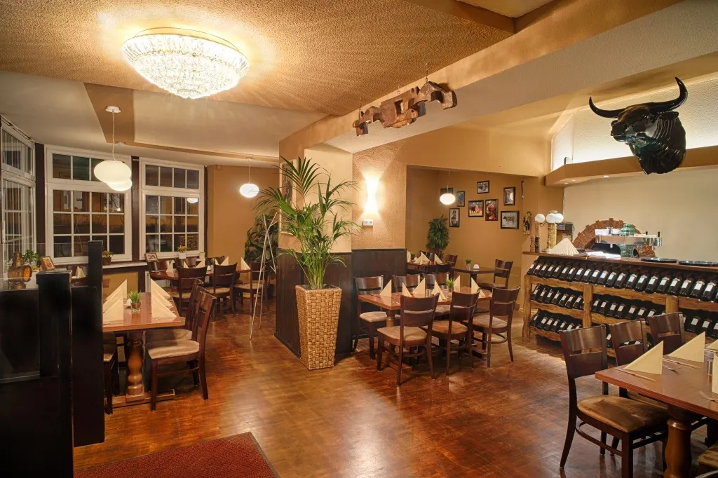 Warm beleuchteter Gastraum mit für Gäste gedeckten Holztischen, gefalteten Servietten, gerahmten Bildern an den Wänden und einem dekorativen Stierkopf, der über der Bar angebracht ist.