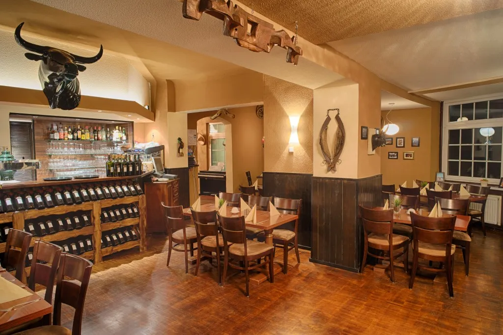 Gemütliches Restaurant-Interieur mit Holztischen und Stühlen, Weinflaschen in Regalen und einem dekorativen Stierkopf an der Wand.
