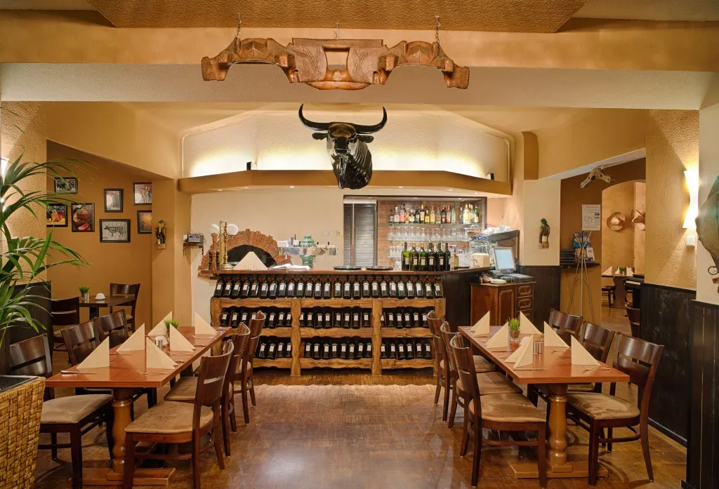 Innenraum eines rustikalen Restaurants mit Holztischen und Stühlen, ordentlich gefalteten Servietten, hinter der Theke ausgestellten Weinflaschen und einem an der Wand montierten Stierkopf.