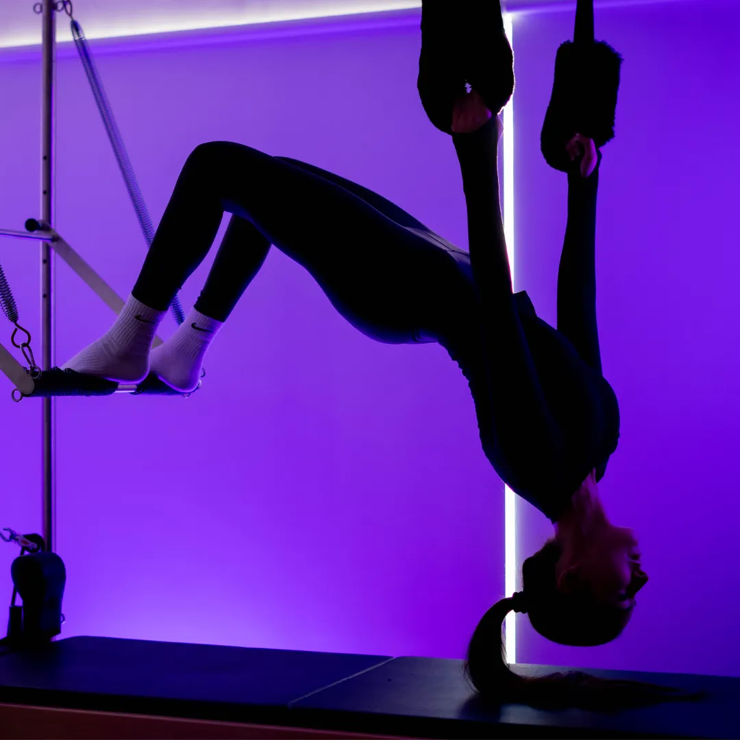 Person performing Pilates exercise on reformer machine against a purple-lit background.
