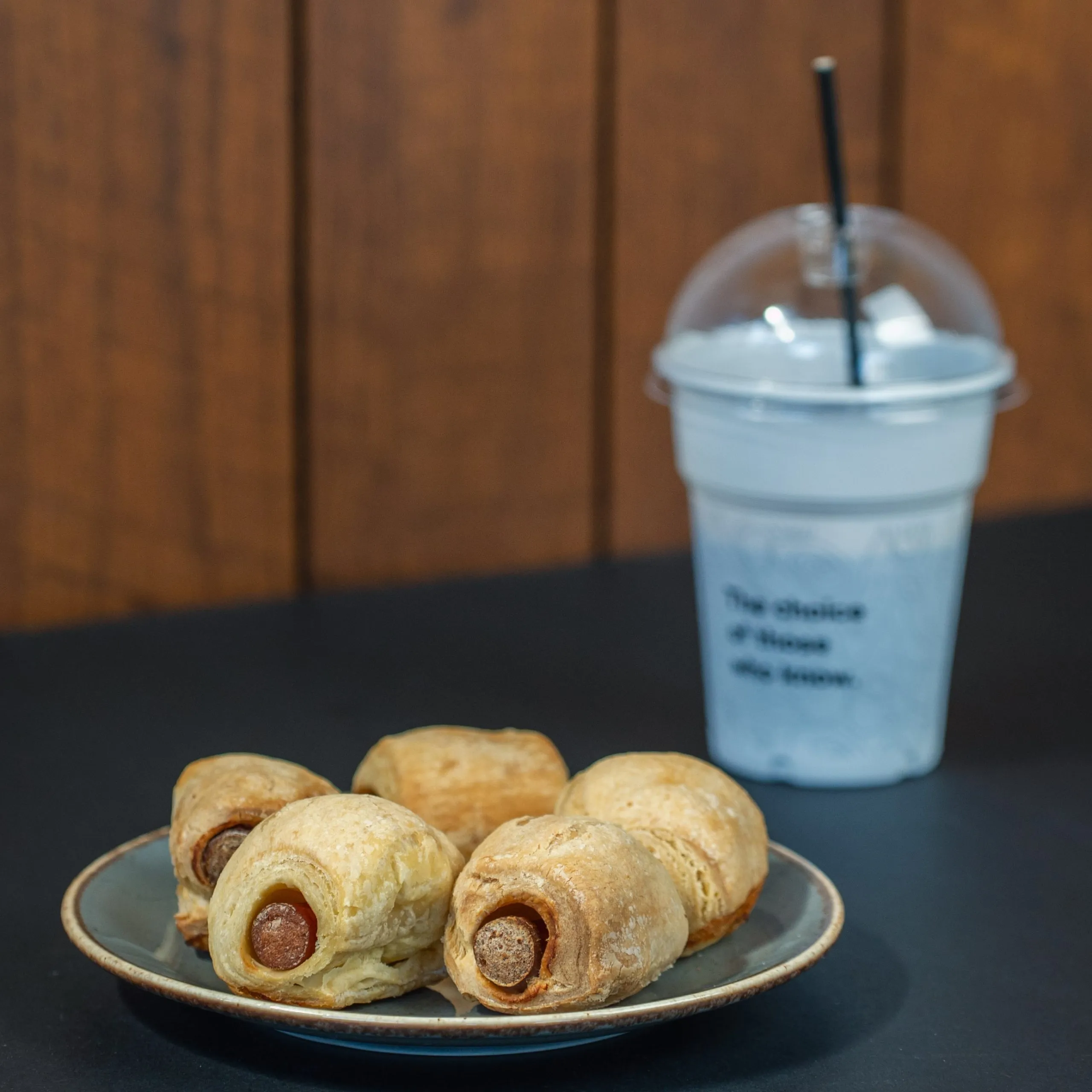 Plate with five small sausage rolls on a dark table next to a plastic cup with a black straw and a dome lid.