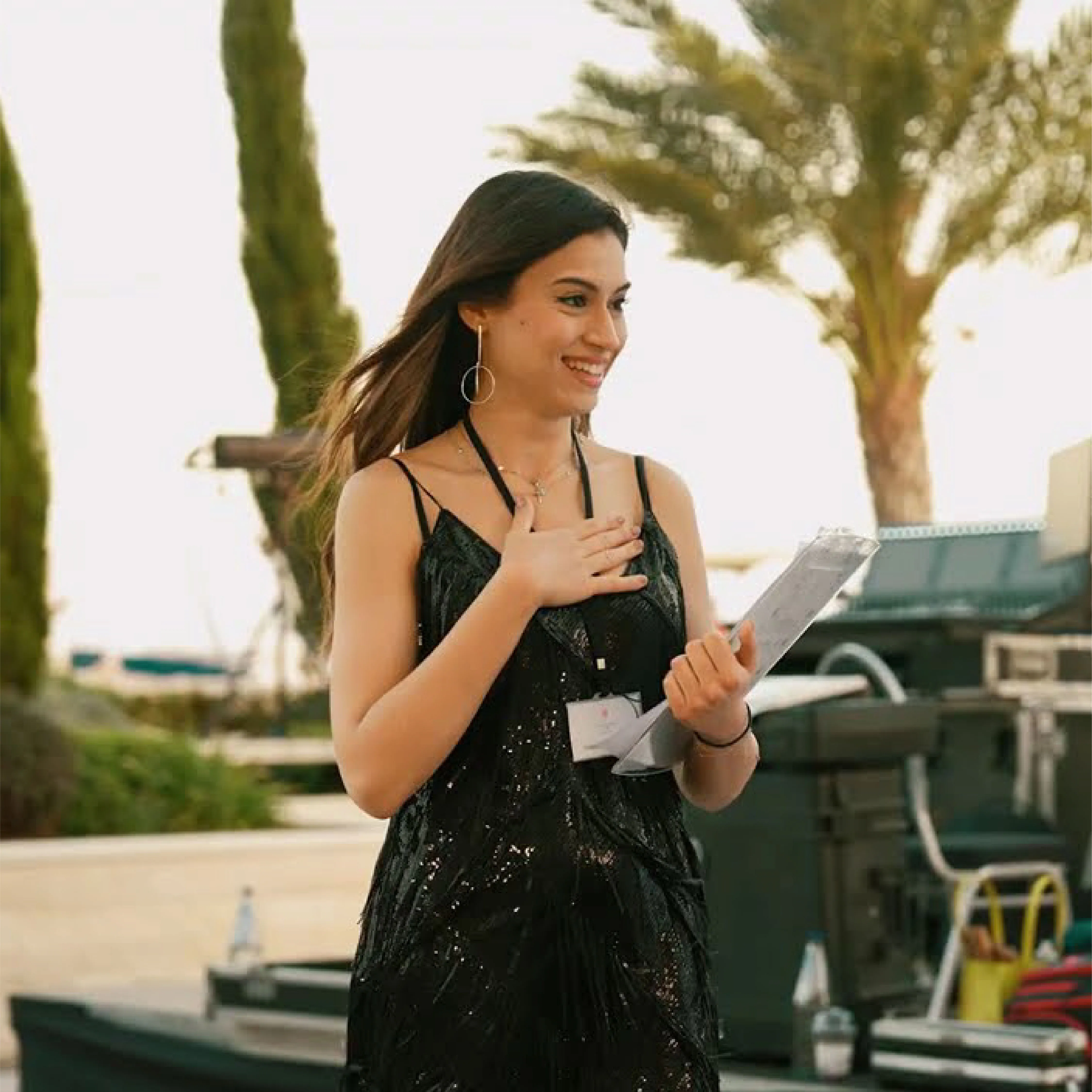Smiling young woman in a black dress holding a wrapped item, with palm trees in the background.