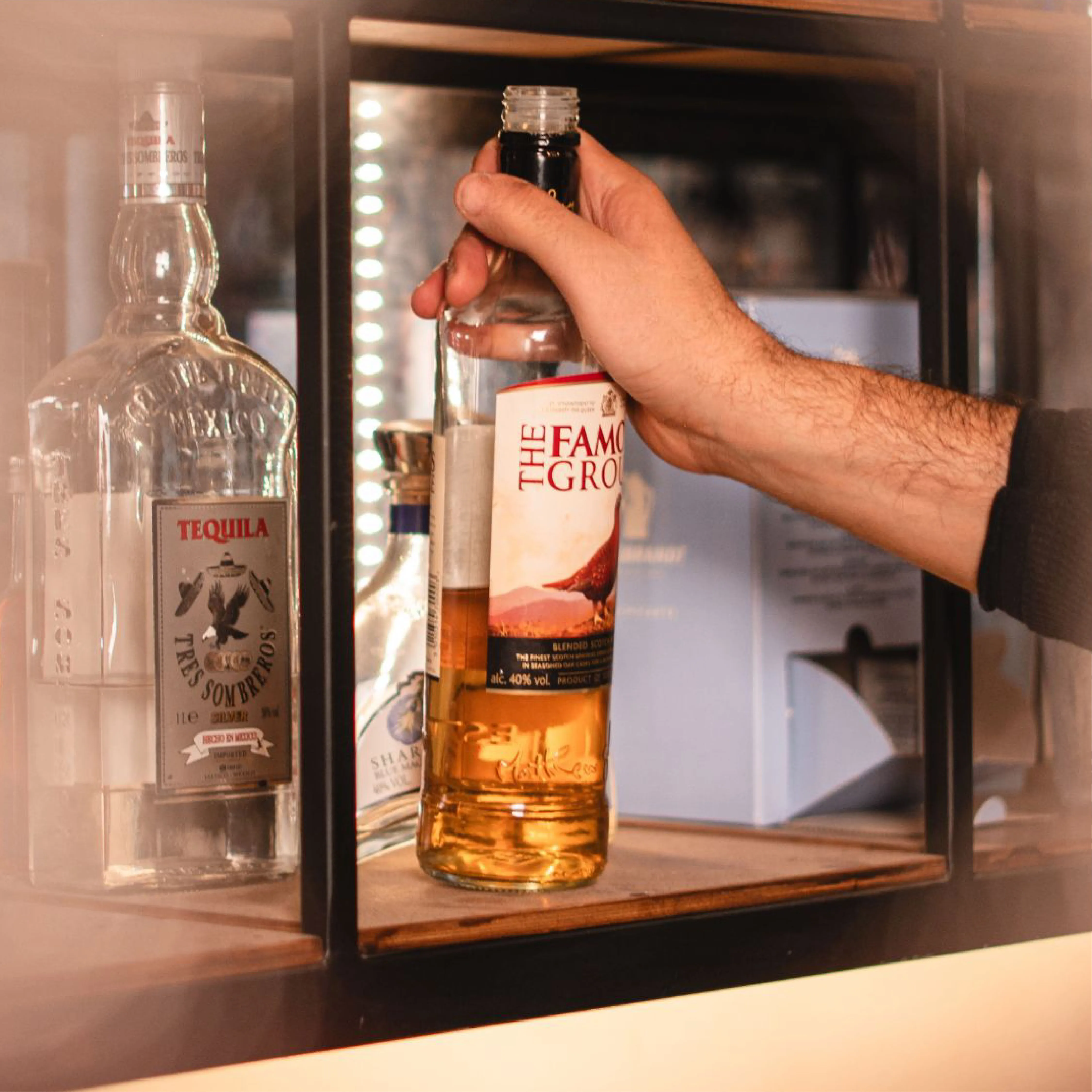 Hand holding a bottle of amber liquor labeled The Famous Grouse on a shelf next to a bottle of Tres Sombreros tequila.