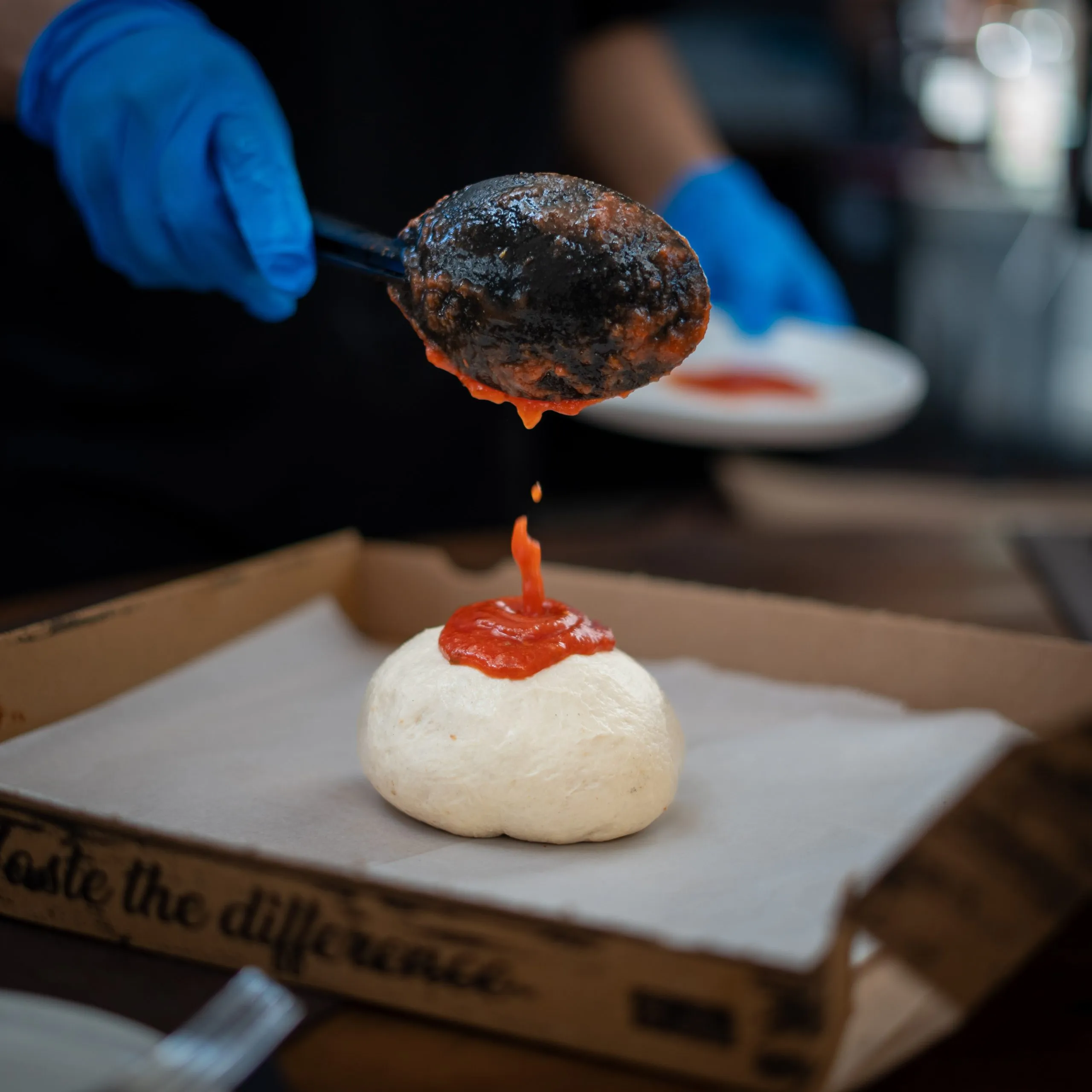 Person wearing blue gloves spooning tomato sauce onto dough ball in a pizza box.
