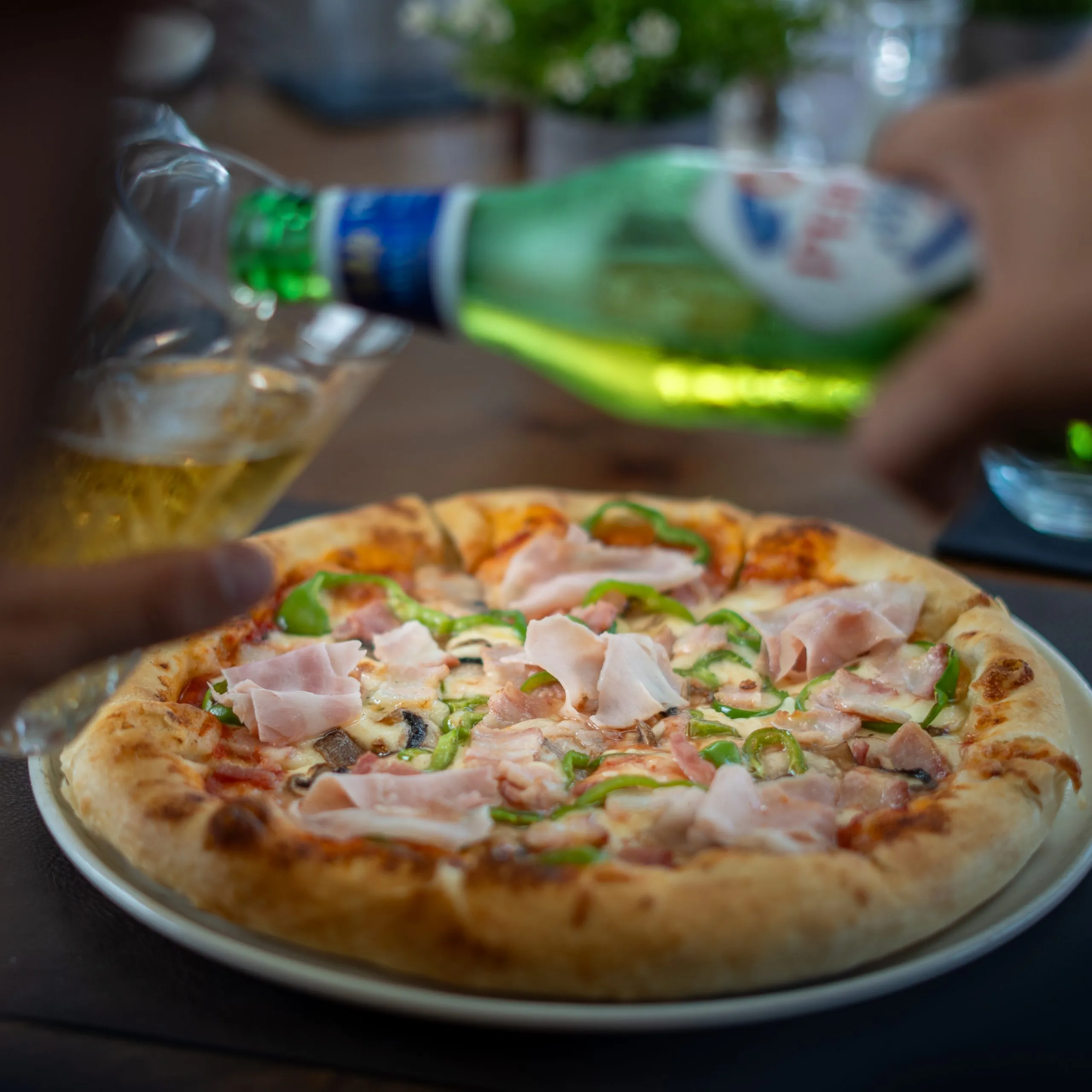 Pizza topped with ham, green bell peppers, and mushrooms on a white plate with a hand pouring beer from a green bottle into a glass.