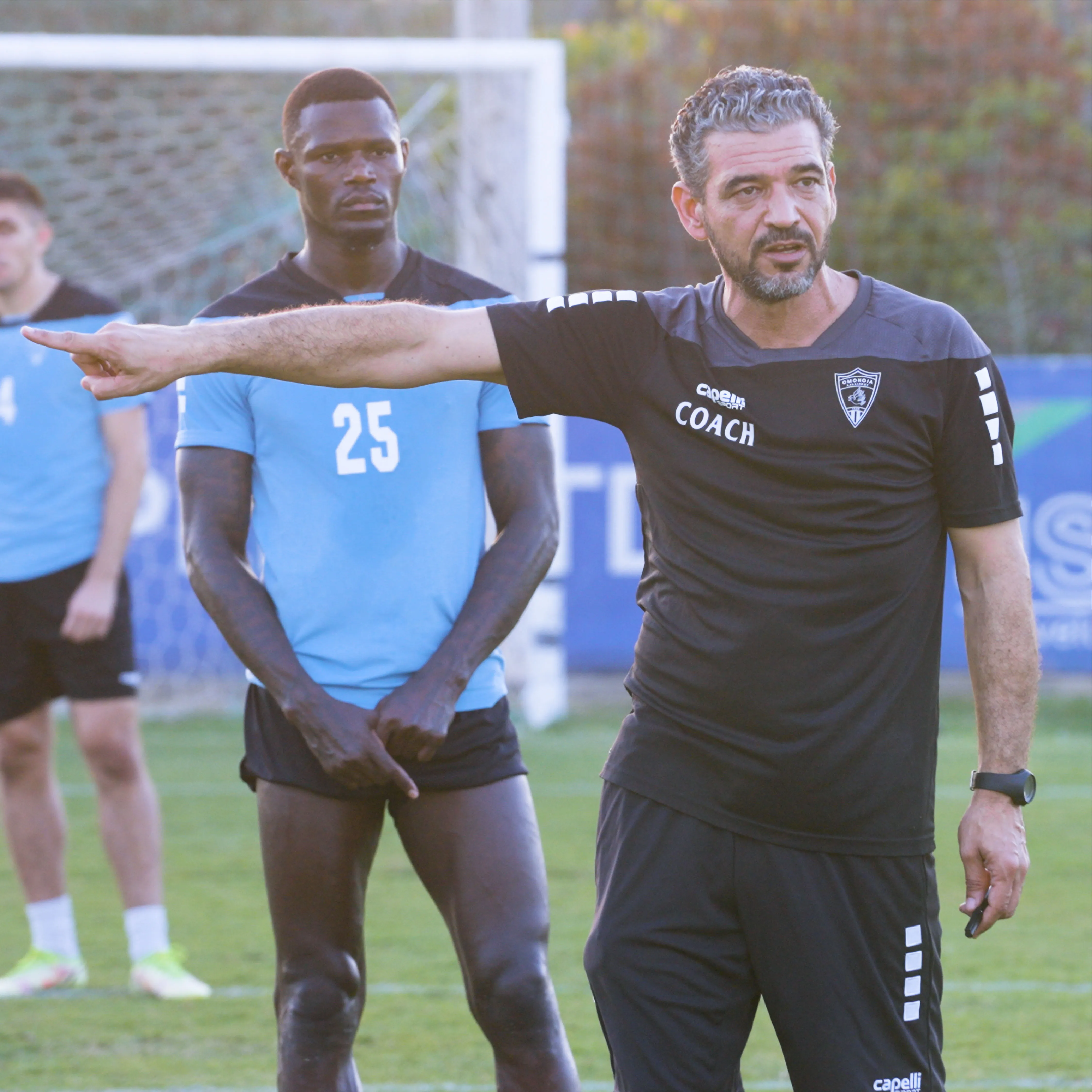 Football coach in black shirt pointing and instructing players wearing light blue jerseys on a grass field with a goal net in the background.