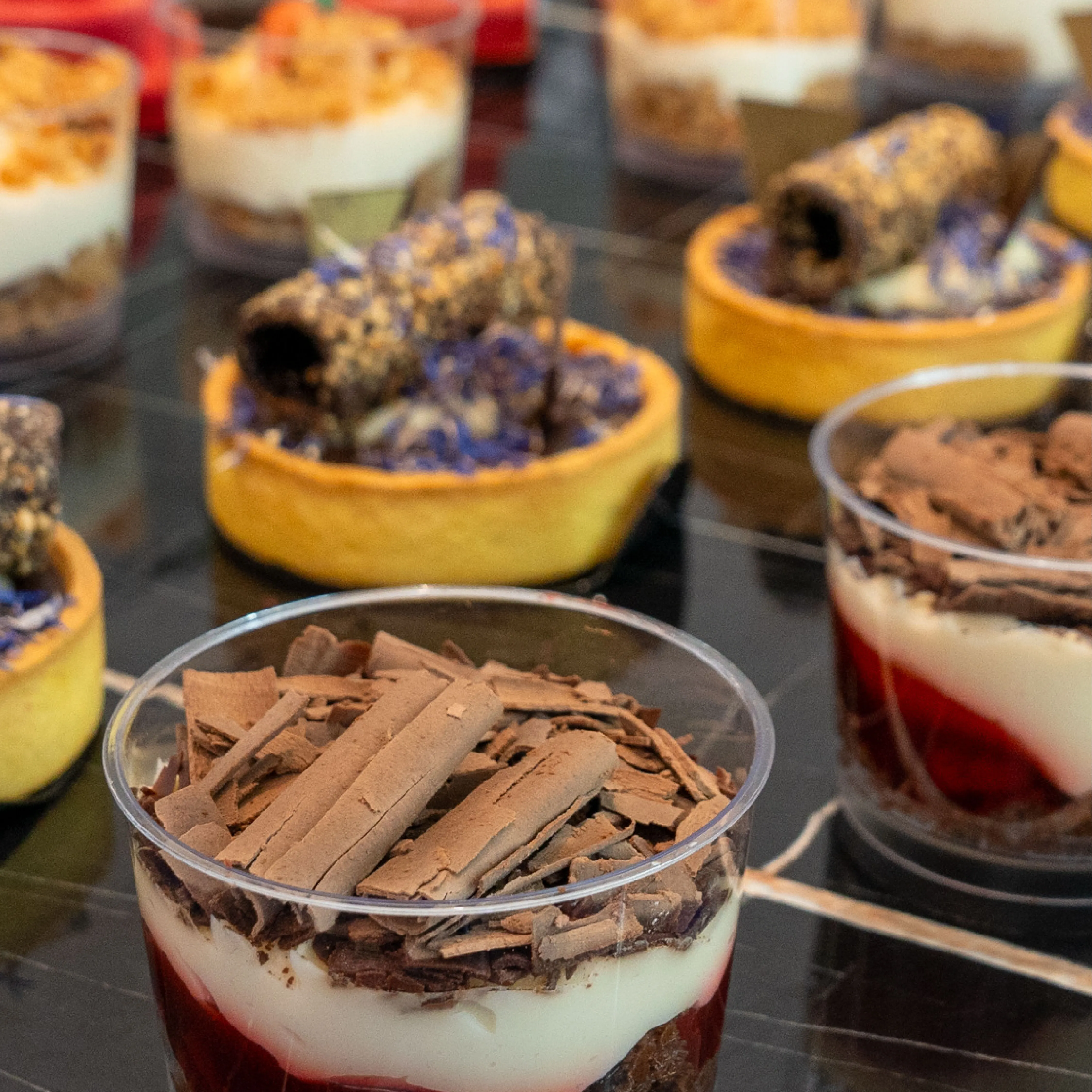 Close-up of layered desserts in clear cups topped with chocolate shavings, with other pastries in the background.