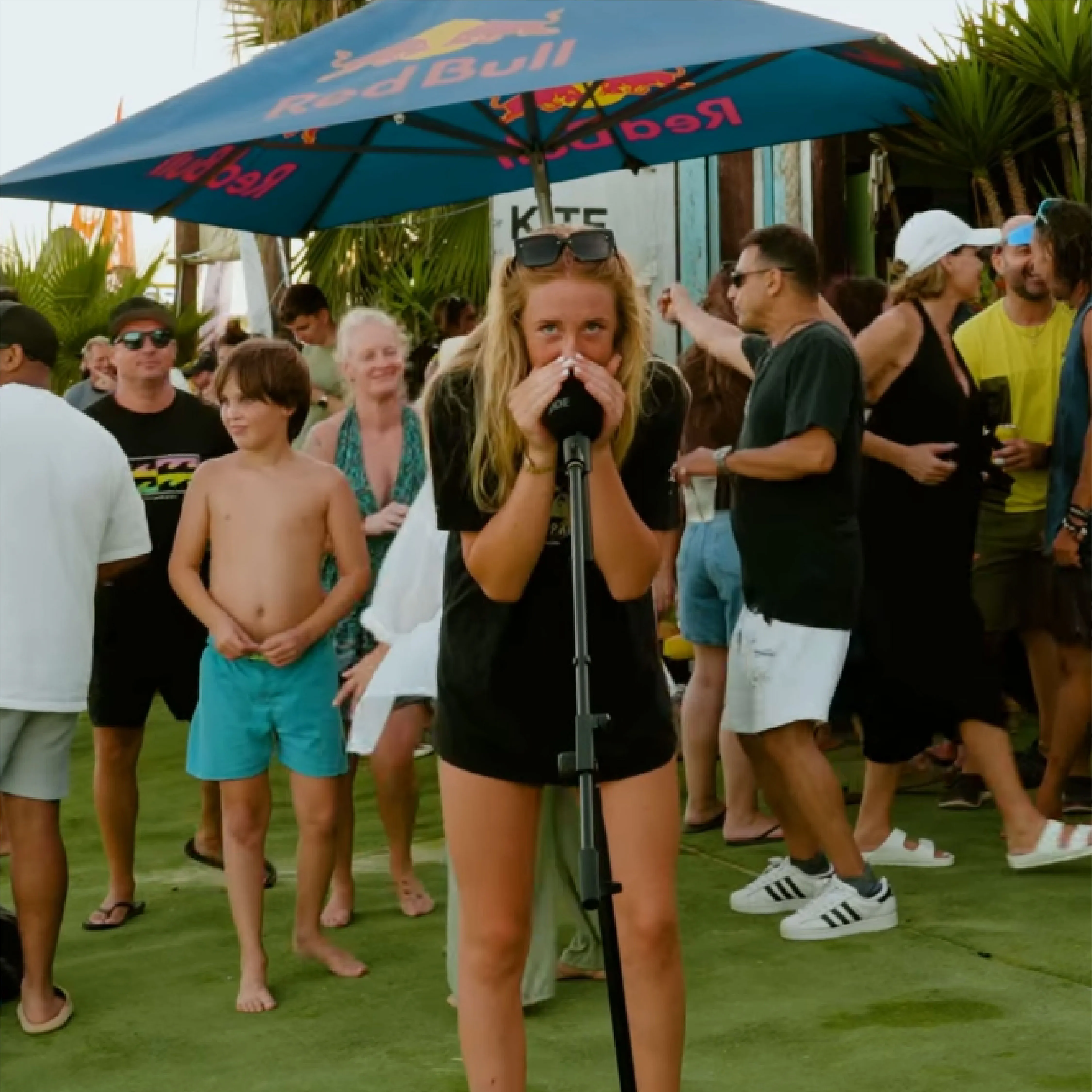 Young woman speaking into a microphone under a Red Bull umbrella at an outdoor event with people socializing in the background.