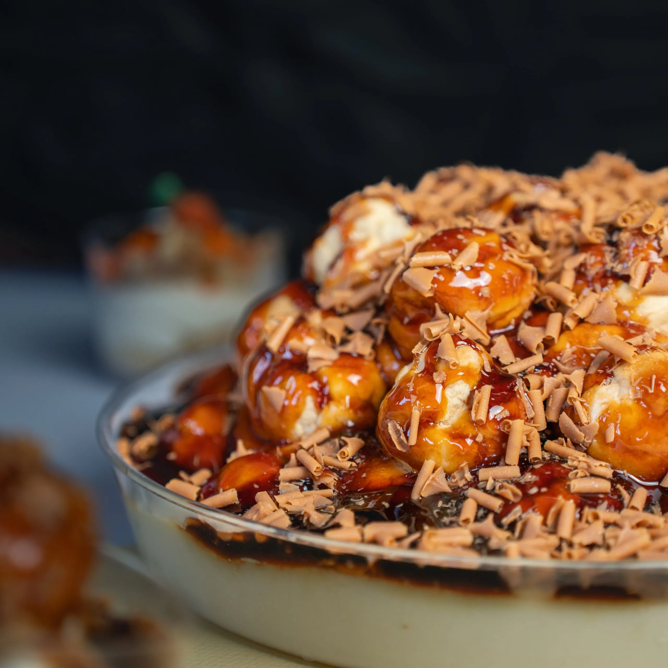 Close-up of a dessert bowl with caramel-coated profiteroles topped with chocolate shavings on creamy base.